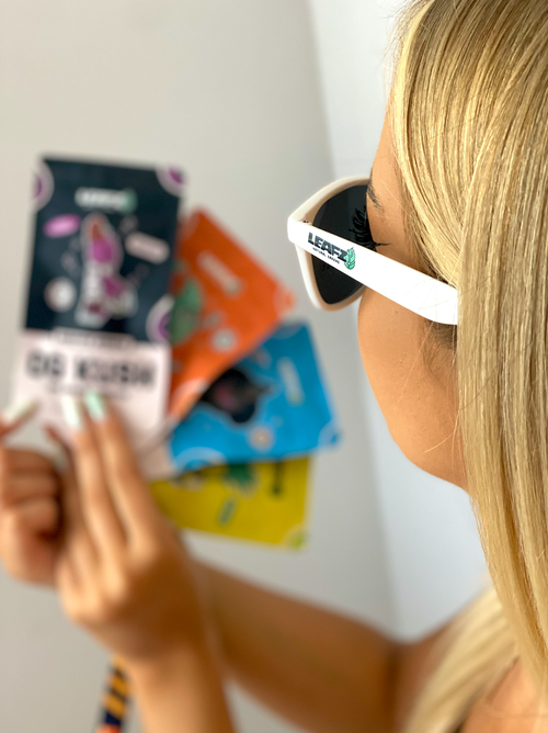 Eine blonde Frau trägt eine weiße Leafz Sonnenbrille und hält vier verschiedene Tüten der HHC Pen in den Händen.